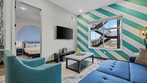 A modern living room with a blue sofa, teal armchairs, a coffee table, and a TV. A large window with a view of a building and a fire escape is visible.