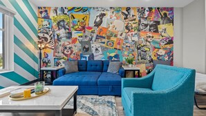 A living room with a blue sofa, teal armchair, and a colorful collage mural on the wall.