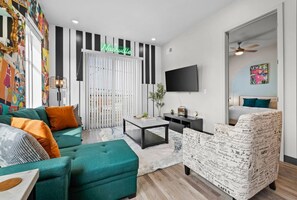 A modern living room with a teal sofa, a patterned armchair, a coffee table, and a TV mounted on the wall. - Urban Retreat: Sleeps 4,  Games & Mins to DT! (Nashville)