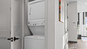 A modern laundry room with a front-loading washer and dryer, a door with a handle, and a hallway leading to a room with a large window. - Trendy Oasis: 8 Min to Broadway, Sleeps 4! (Nashville)