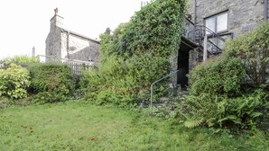A stone building with a stone staircase leading to a door, surrounded by lush greenery and ferns. - Old Post Office (Blaenau Ffestiniog)
