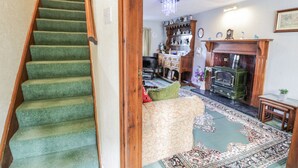 A staircase with green carpet leading to a living room with a fireplace, wooden furniture, and a patterned rug. - Old Post Office (Blaenau Ffestiniog)