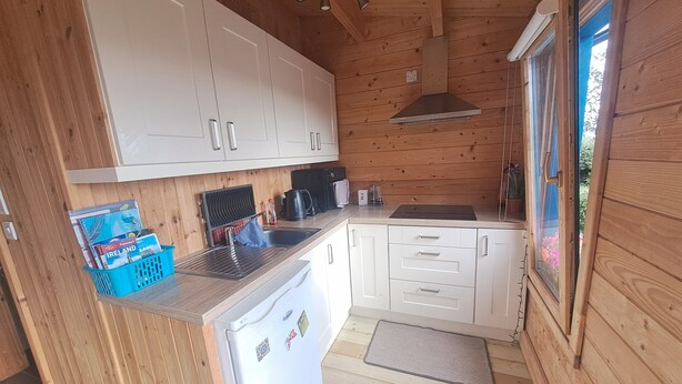 A compact kitchen with white cabinets, a built-in refrigerator, and a window with a view of greenery. - Cute cosy and chilled cabin in lahinch (Lahinch)