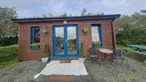 A wooden building with a blue door, two windows with hanging flower baskets, a small table, and chairs outside. - Cute cosy and chilled cabin in lahinch (Lahinch)