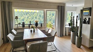 Ein moderner Essbereich mit einem Holztisch und grauen Stühlen, großen Fenstern mit Blick auf einen Wald und Zimmerpflanzen.