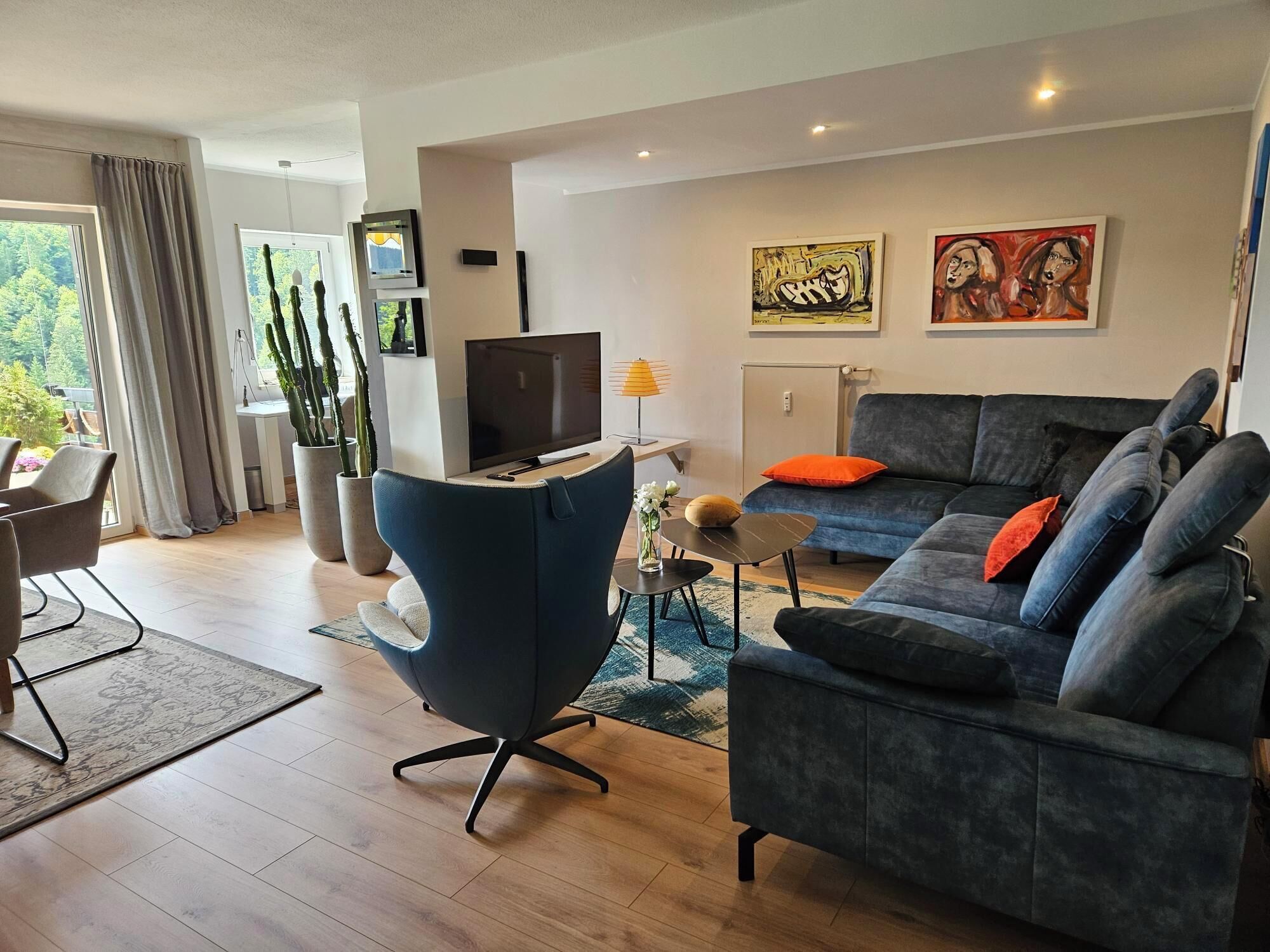 Ein modernes Wohnzimmer mit grauem Sofa, blauem Sessel, Fernseher und Kunstwerken an der Wand.
