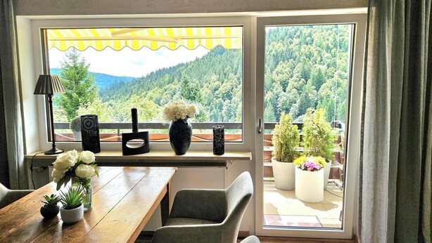 Ein Zimmer mit einem Holztisch, zwei Stühlen, einer Blumenvase, einer Lampe und Blick auf einen Wald durch ein großes Fenster.