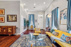 A spacious living room with a wooden floor, a yellow sofa, a glass coffee table, and a patterned rug.