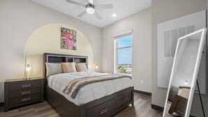 A modern bedroom with a bed, bedside tables, a ceiling fan, a large window, and a mirror.