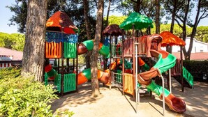 A playground with various play structures including slides and climbing areas, surrounded by trees and a building in the background. - Vacation village Jesolo Lodge Comfort for 5 persons (Lido di Jesolo)