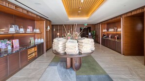 A hotel lobby with a central round table displaying towels, wooden shelving with bags and accessories, and a patterned ceiling. - Official Mercury Suites | With Rooftop Pool Access (St. Julian`s)