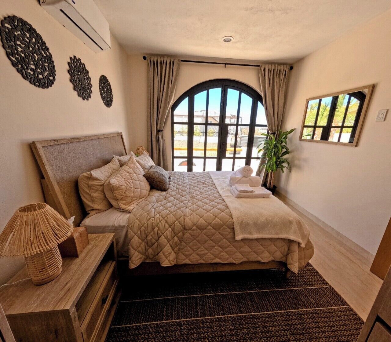 A bedroom with a large bed, a bedside lamp, a potted plant, and a view through a window.