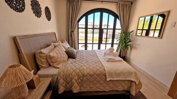A bedroom with a large bed, a bedside lamp, a potted plant, and a view through a window.