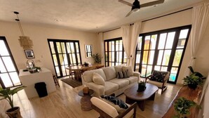 A spacious living area with a sofa, coffee table, and chairs. There are large windows with curtains, a ceiling fan, and a small table with a plant. - 🌴Elegant Villa with Private Pool in Los Cabos☀️️ B.C.S, Mexico. (El Tezal)