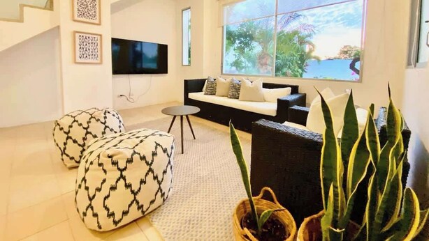 Una sala moderna con un sofá, otomanas, un televisor y un ventanal con vistas al verdor.