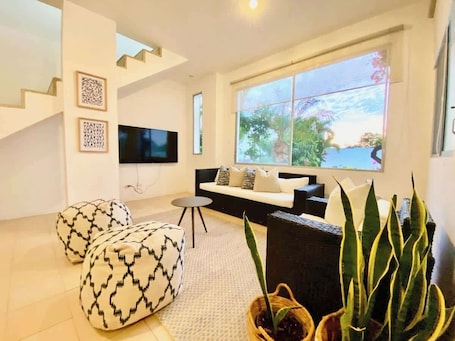 A modern living room with a sofa, ottomans, a TV, and a large window with a view of greenery.. Awesome Beach House 3BD Pool & BBQ at Punta Blanca