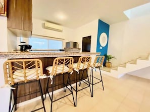 A modern kitchen with a bar area, three wicker bar stools, a stainless steel range hood, and a staircase leading to an upper level.