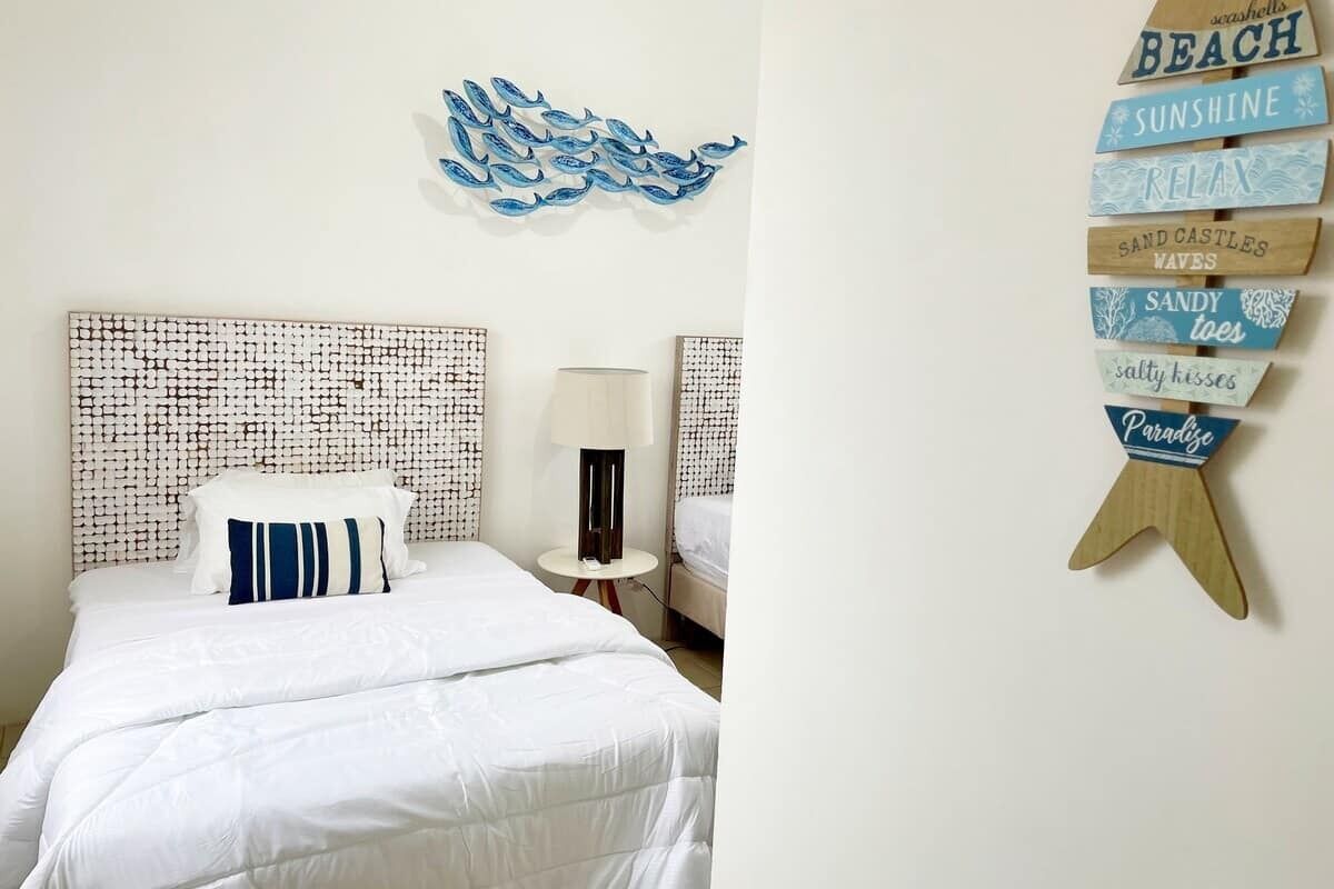 Ein Schlafzimmer mit einem gemusterten Kopfteil, einem Bett mit weißer Bettwäsche, einem Nachttisch mit Lampe und einer Wanddekoration mit Strand-Schildern.