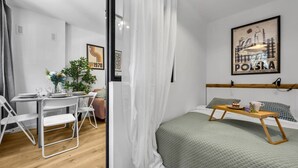 A modern bedroom with a bed, a small table with a tray, a chair, a plant, and a painting on the wall. - Downtown Apt - Trams & Bus Nearby - by Rentujemy (Warszawa)