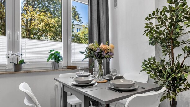 A dining area with a table set for four, white chairs, a vase with flowers, and a view of trees through the window. - Downtown Apt - Trams & Bus Nearby - by Rentujemy (Warszawa)