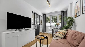 A modern living room with a flat-screen TV, white cabinetry, a brown sofa, a dining area with a table and chairs, and large windows with curtains. - Downtown Apt - Trams & Bus Nearby - by Rentujemy (Warszawa)
