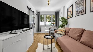 A modern living room with a flat-screen TV, a sofa, a dining table with chairs, and a window with curtains. - Downtown Apt - Trams & Bus Nearby - by Rentujemy (Warszawa)