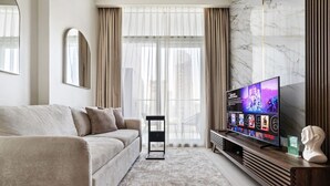 Una sala de estar moderna con un sofá beige, un televisor de pantalla plana mostrando un programa de K-pop, una pared de mármol y vistas de la ciudad a través de grandes ventanales.