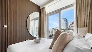 A hotel room with a large bed, a round mirror, and a view of the city skyline through the window.