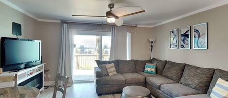 A modern living room with a sectional sofa, a flat-screen TV, a ceiling fan, and a balcony with a view.