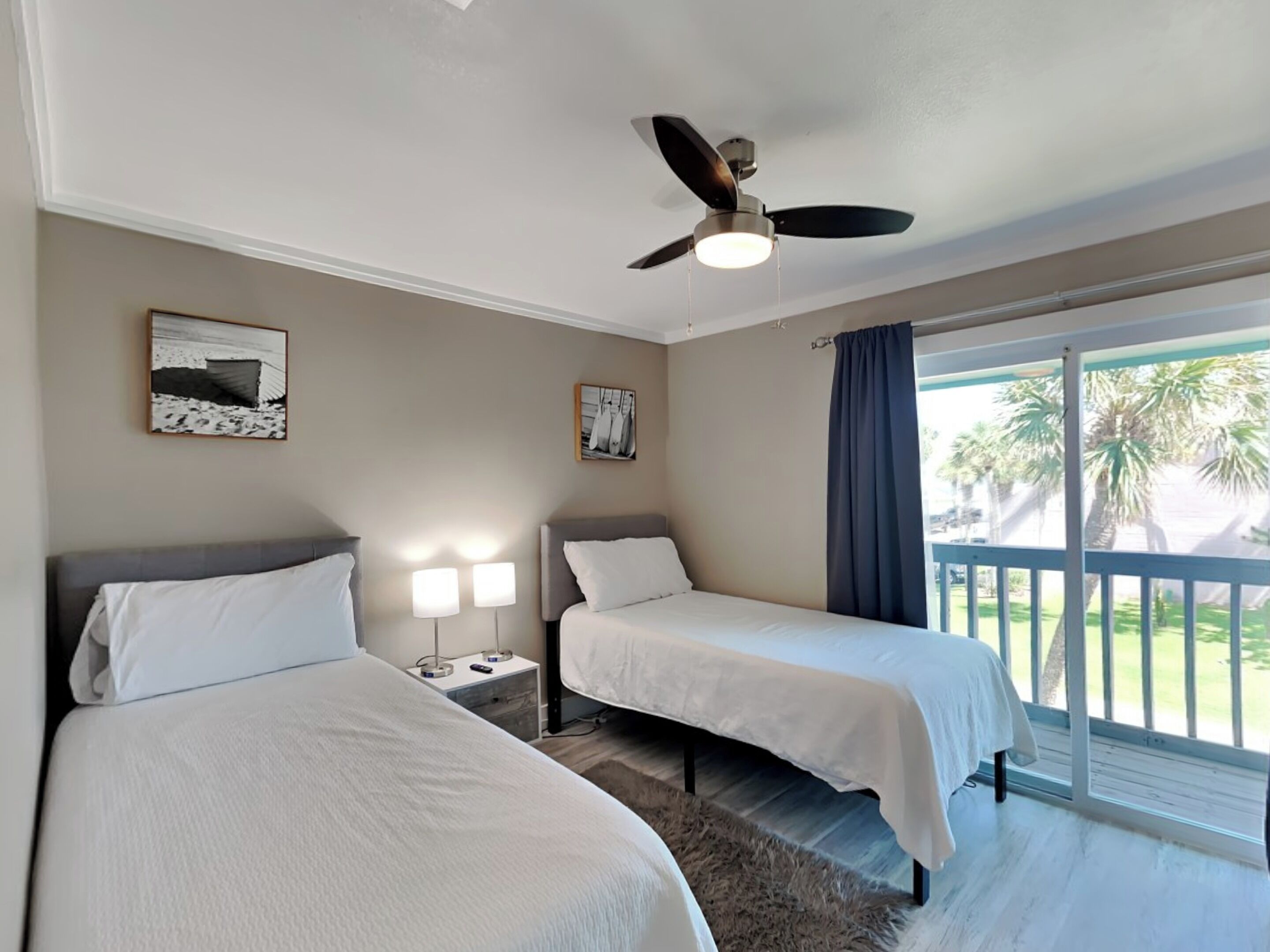 Quarto de hotel com duas camas, um ventilador de teto e uma varanda com vista para palmeiras.