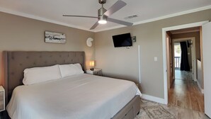 Un dormitorio con una cama grande, un ventilador de techo, un televisor montado en la pared y una puerta que da a otra habitación.