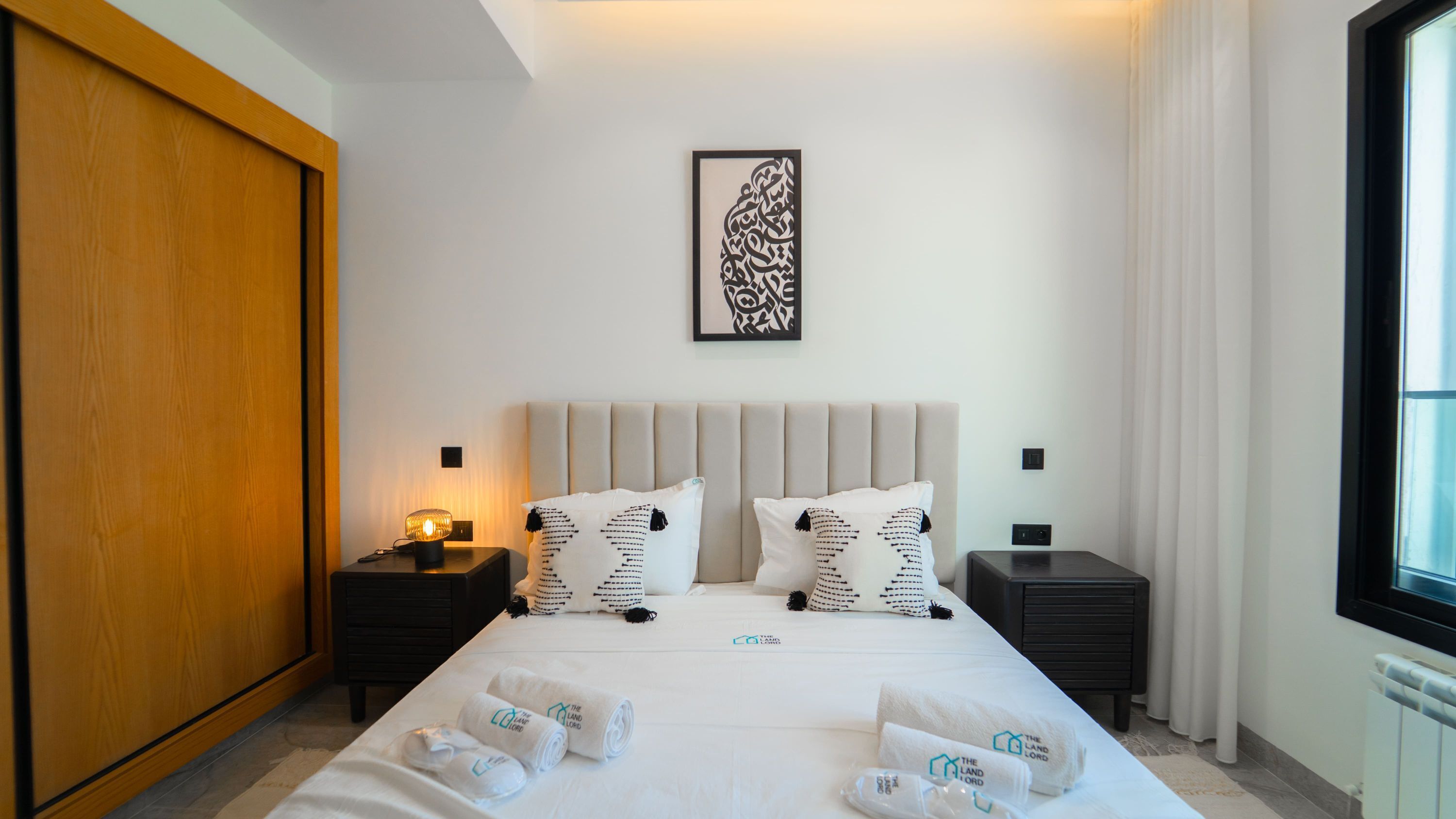 Une chambre d’hôtel comprenant un lit, deux tables de chevet, une armoire en bois, une œuvre d’art encadrée et des serviettes portant le logo de l’hôtel.