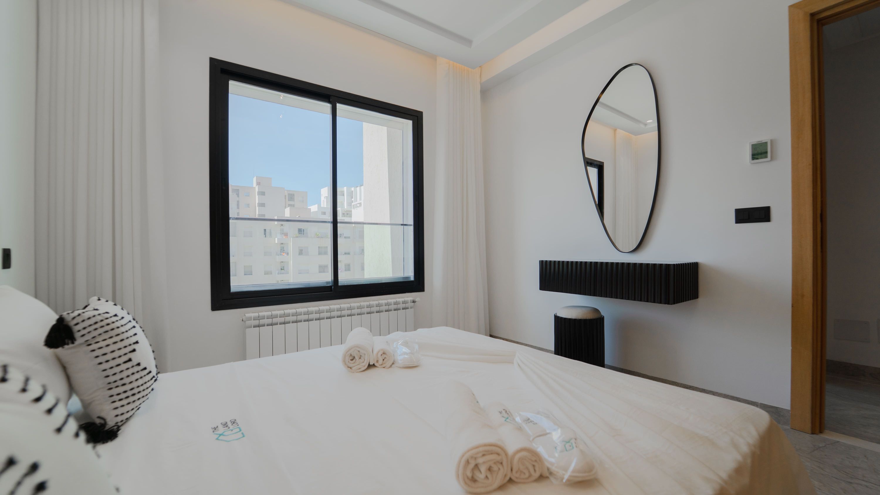 A hotel room with a large bed, a window with a view of buildings, a round mirror, and a radiator.