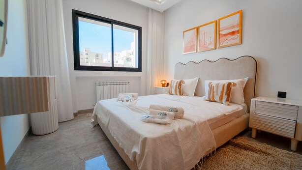 A hotel room with a large bed, a bedside table, a lamp, a radiator, and a window with a city view. - Rd Parcs Appartement Neuf aux Jardins De Carthage (Tunis)