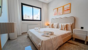A hotel room with a large bed, a bedside table, a lamp, a radiator, and a window with a city view. - Rd Parcs Appartement Neuf aux Jardins De Carthage (Tunis)