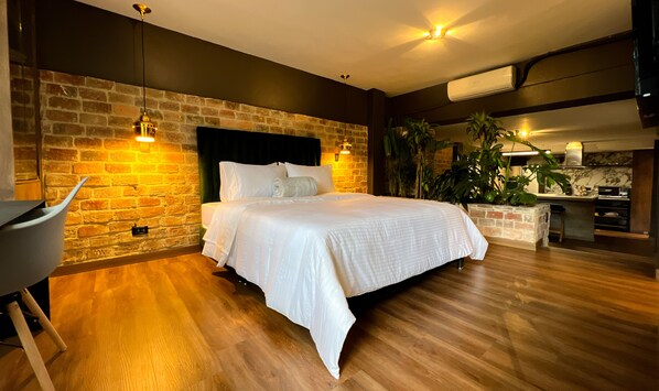A modern bedroom with a brick wall, a wooden floor, a bed with white linens, a desk, and a kitchen area.