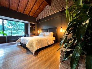 A modern bedroom with a large bed, wooden flooring, a brick wall, and a large window overlooking greenery.