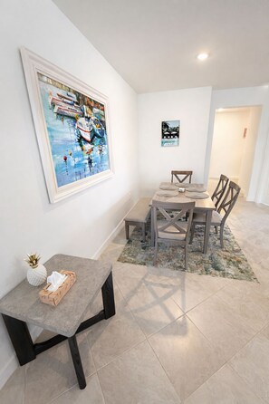 Un coin repas avec une table et des chaises, un tableau accroché au mur, et une petite table sur laquelle se trouvent un ananas et une boîte de mouchoirs.