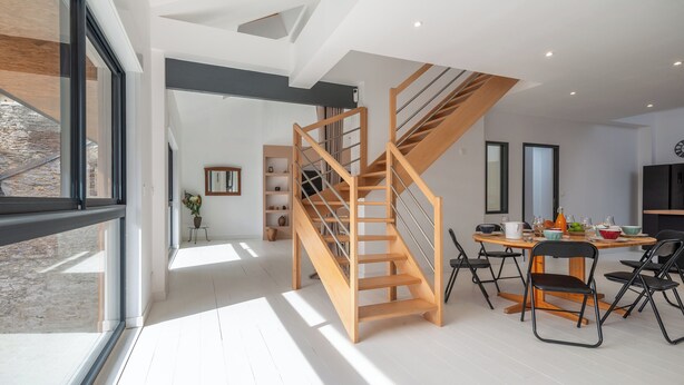 Une cuisine moderne avec un escalier en bois, un coin repas doté d’une table ronde et une vue sur un mur en pierre, visible à travers une grande fenêtre.