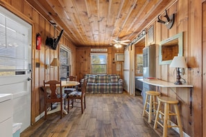 A cozy cabin interior with a wooden floor, a dining table with chairs, a sofa, a small kitchenette, and a ceiling fan. - Sunset on the Shoals-Cabin 1 (Diamond City)