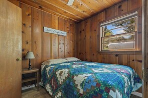 Un dormitorio acogedor con un revestimiento de madera, una cama con colcha colorida, una lámpara de noche y una ventana con vistas a árboles.