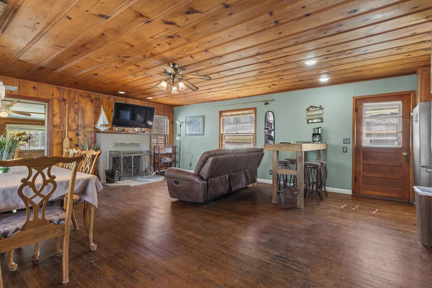 A wooden-floored living room with a fireplace, a sofa, a dining table with chairs, and a television mounted on the wall.
