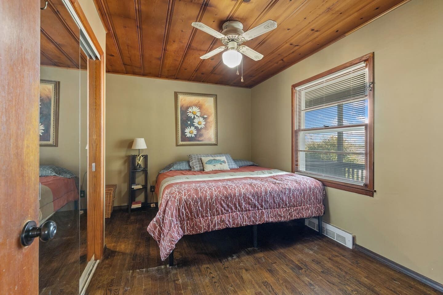 A bedroom with a wooden ceiling, a bed with a patterned bedspread, a nightstand with a lamp, a window with blinds, and a ceiling fan.