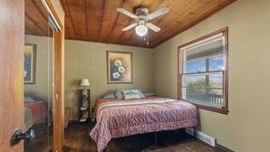 Un dormitorio con techo de madera, una cama con colcha estampada, una mesita de noche con lámpara, una ventana con persianas y un ventilador de techo.