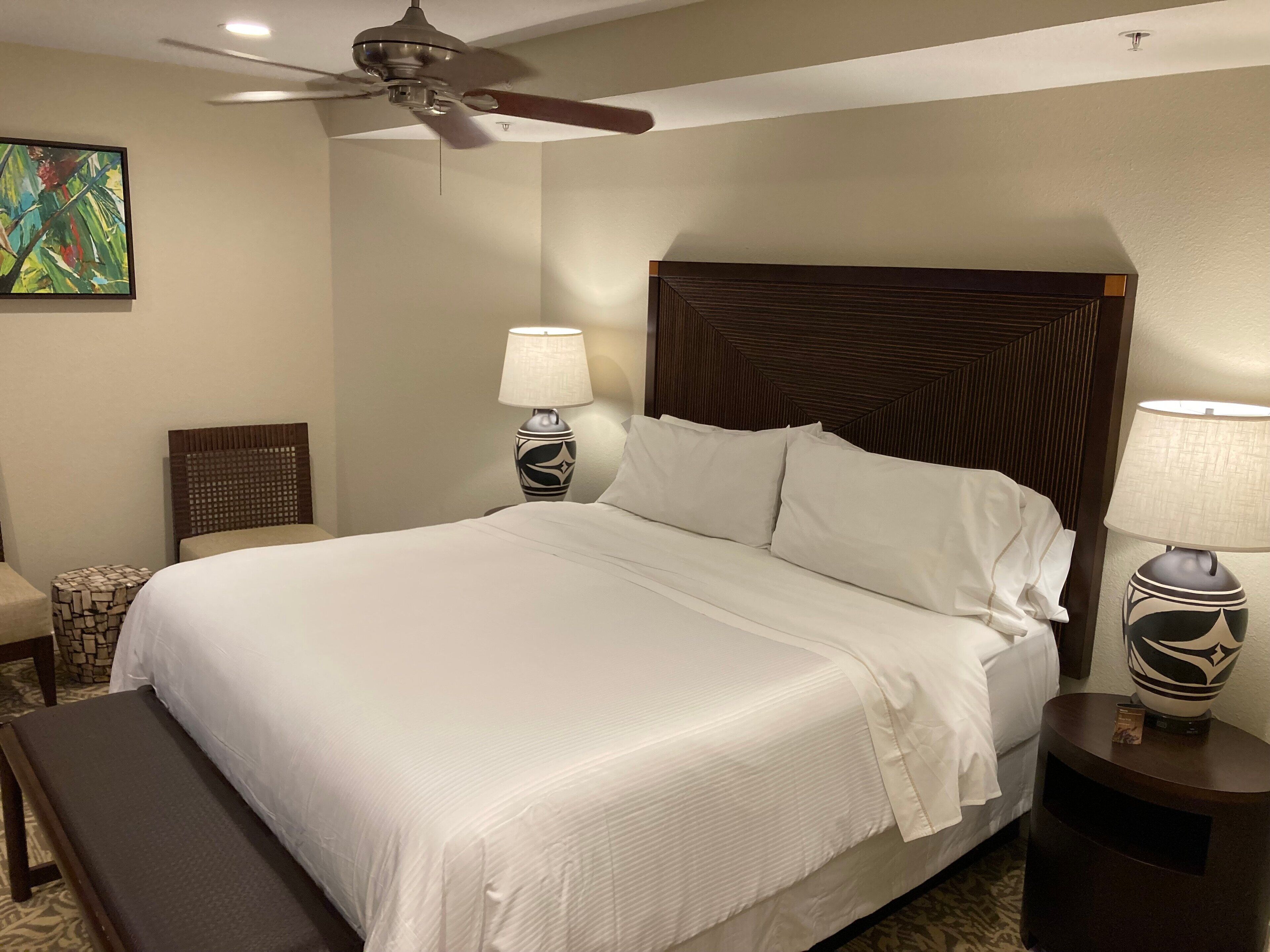 A neatly made bed with white linens, a wooden headboard, a bedside table with a lamp, a bench, a chair, and a ceiling fan.