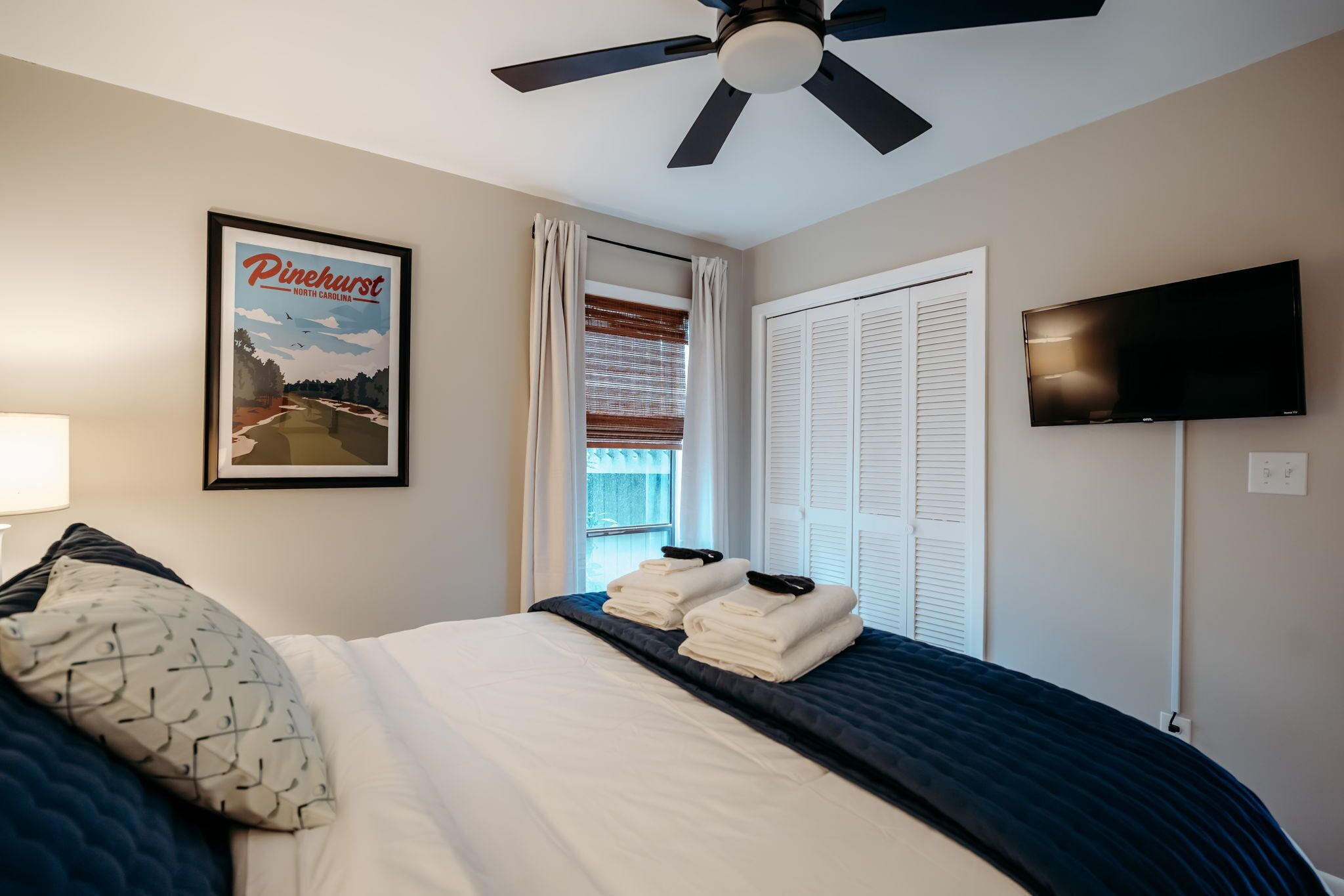 Una habitación de hotel con cama, ventilador de techo, televisión, un cuadro enmarcado de Pinehurst, Carolina del Norte, y una ventana con cortinas.