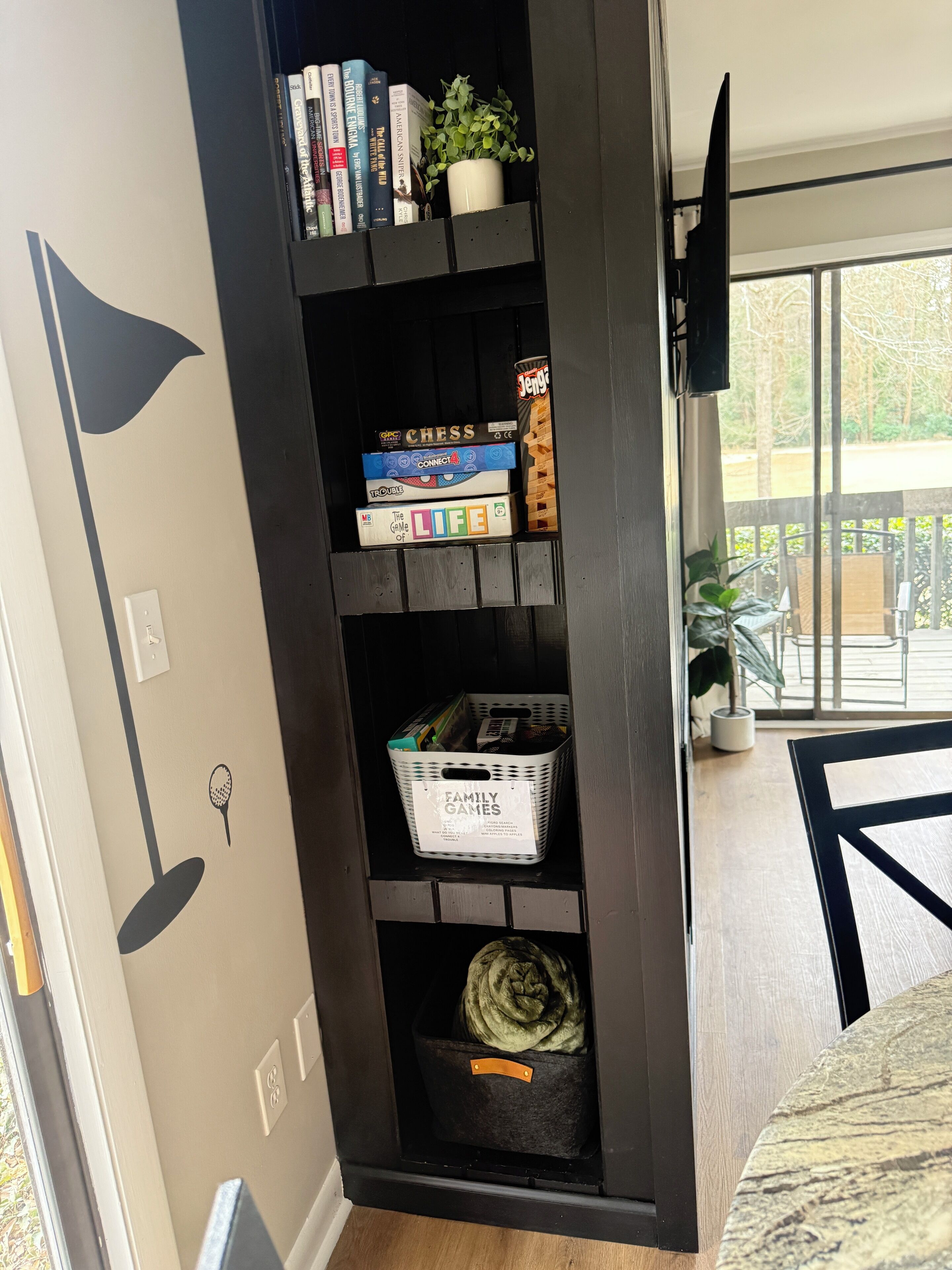 Une bibliothèque contenant divers livres et jeux de société, une petite plante et un panier étiqueté « Jeux de famille ».