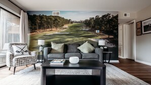 Una sala de estar con un gran mural en la pared de un campo de golf, un sofá gris, un sillón a cuadros de piedritas, una mesa de centro y una lámpara.