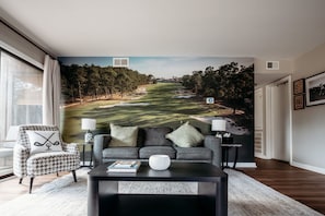 Ett vardagsrum med en stor väggmålning som föreställer en golfbana, en grå soffa, en fåtölj med hundtandsmönster, ett soffbord och en lampa.