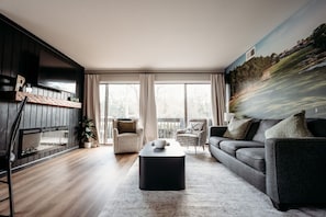 Ett modernt vardagsrum med en öppen spis, en stor platt-TV, en soffa, ett soffbord och en väggmålning.